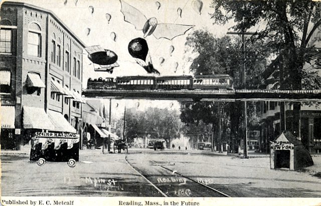 A scan of a black and white postcard which depicts what Reading, Massachusetts might look like in the future. The foreground shows a wide street lined with trees on the right and a three story brick building on the left. Halfway up the postcard is a monorail track with four train vehicles on it traveling to the left. Above that are a dirigible, a hot air balloon, and a plane with bat like wings. Surrounding these items in the sky are multiple smaller hot air balloons.