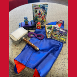 Blue satin cape, purple mask, foam mallet, three wooden superhero dolls and 3 superhero books arranged on a gray ottoman